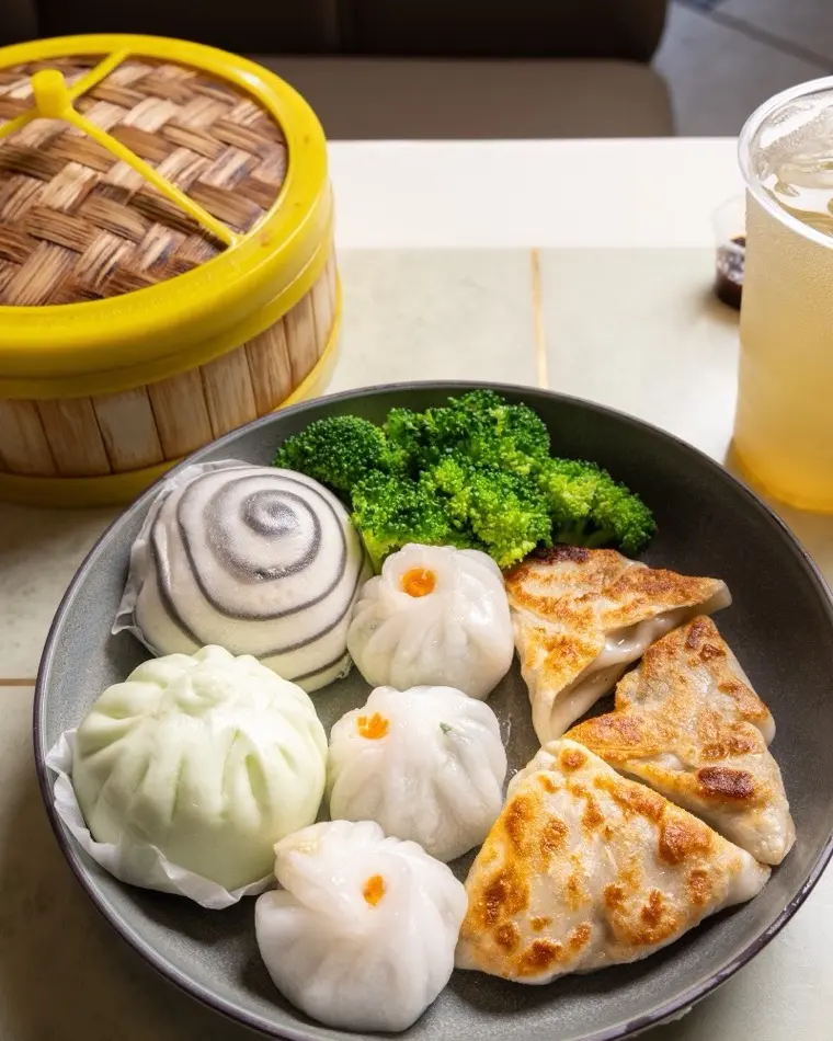 Steamed bao buns, pork siu mai and golden pan-fried pot stickers with broccoli on a dark plate