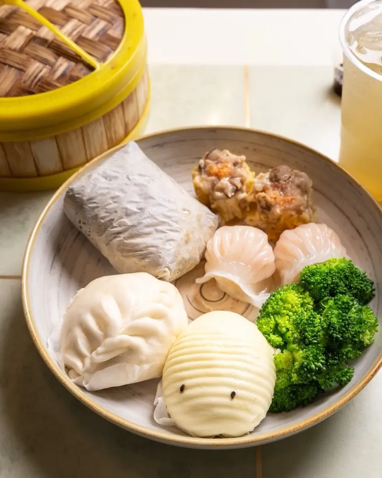 Dim sum sampler with shrimp har gow, pork siu mai, a sticky rice wrap, steamed bun and broccoli beside a bamboo steamer