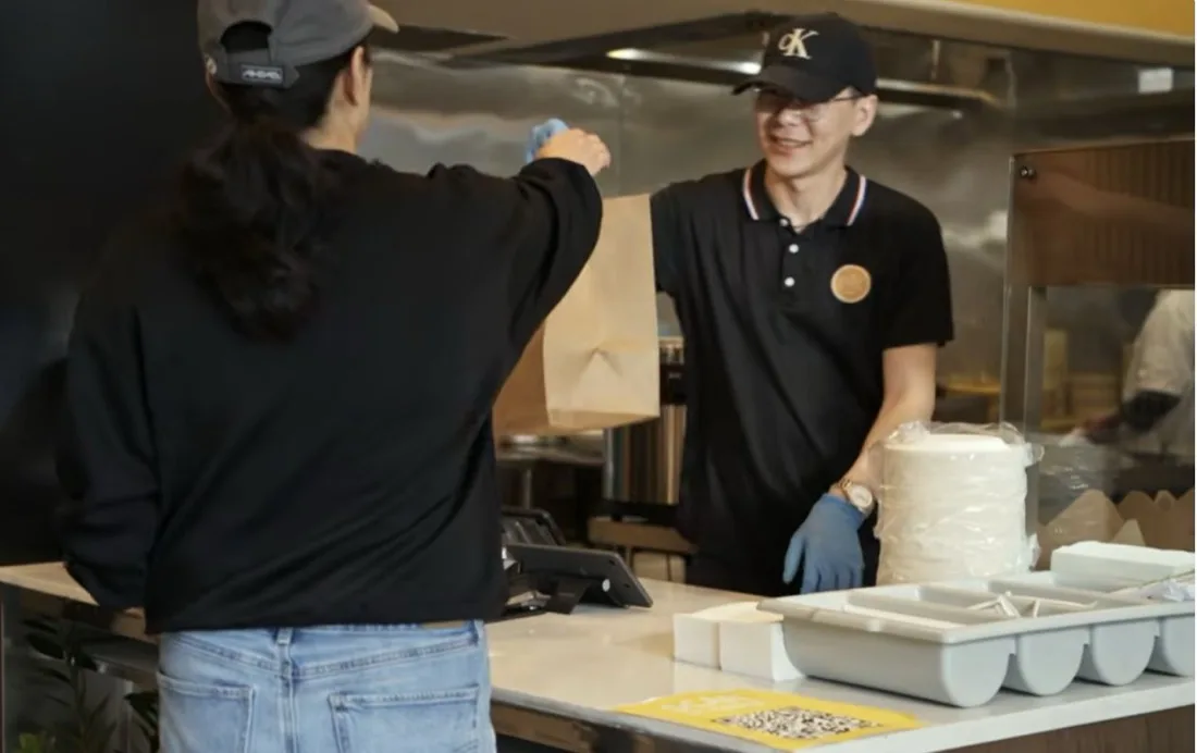A smiling Bao Express team member handing a fresh takeout order across the pickup counter to a guest